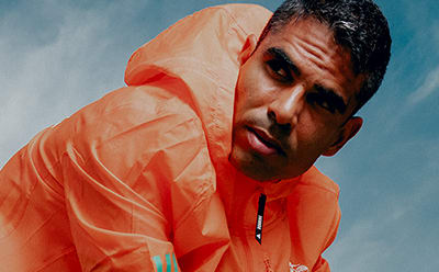A close-up portrait of a man wearing a bright orange shirt against a clear blue sky background.