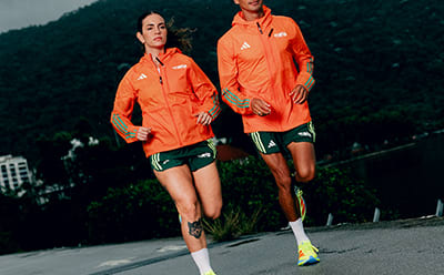 Two individuals wearing bright orange athletic jackets and shorts are running on a paved path surrounded by lush, green foliage in the background.
