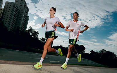 Two runners in athletic gear are jogging on a paved path surrounded by tall buildings and palm trees, with a bright blue sky overhead.