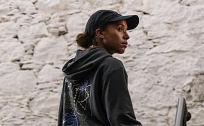 A young person wearing a black hooded sweatshirt and cap stands in front of a weathered stone wall.