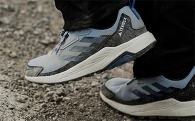 The image shows a pair of gray and blue athletic shoes with a distinctive treaded sole, standing on a gravel or rocky surface.