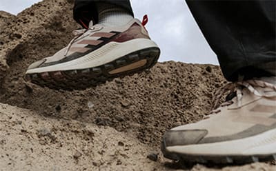The image shows a pair of hiking boots or sneakers with a rugged, textured sole standing on a sandy, rocky surface in the foreground, with a blurred background suggesting an outdoor, mountainous environment.