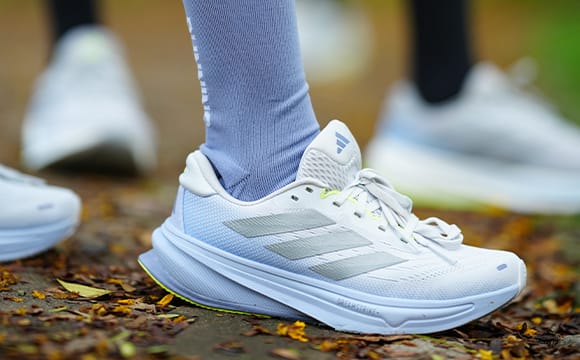 A pair of white and gray athletic shoes with thick soles stand on a forest floor covered in fallen leaves, with blurred trees and foliage in the background.