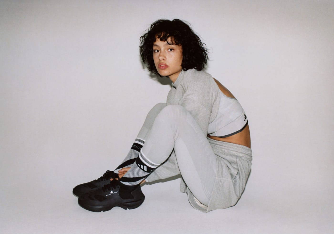 A model sitting on the floor. She wears a layered look of the cropped sweatshirt with cropped T-shirt underneath on top. On bottom she wears the grey tight underneath the Terry shorts.