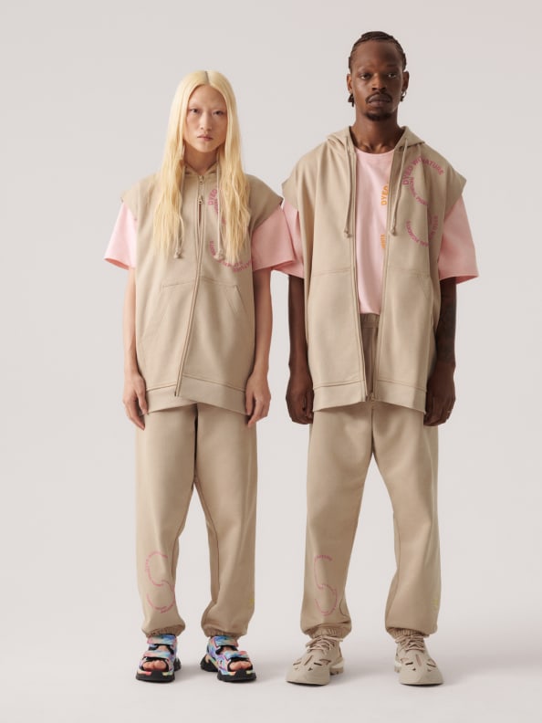 Two models standing, they wear the new sleeveless hoodie and sweat pants from the SS23 adidas by Stella McCartney gender-neutral loungewear drop. Crafted from 100% organic cotton.