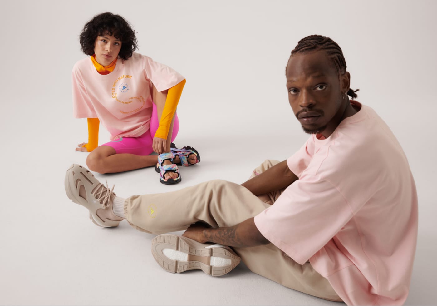Two models sitting, they wear the new SS23 adidas by Stella McCartney gender-neutral loungewear drop, crafted from 100% organic cotton.