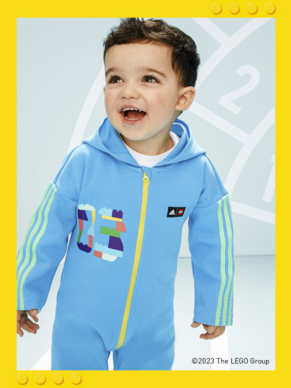 A infant boy standing in front of a printed athletics track, smiling playfully, wearing adidas LEGO® onesie.