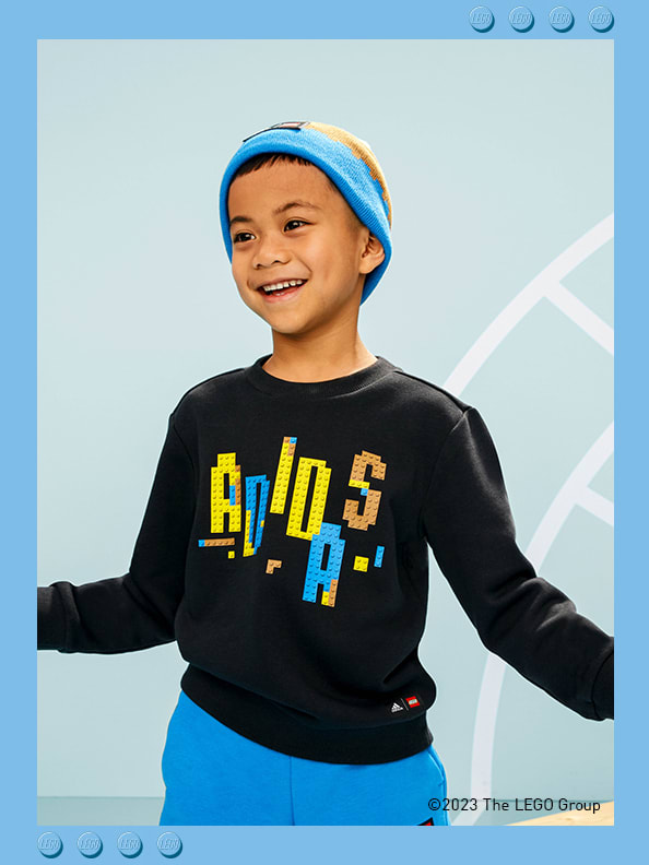 A little boy standing in front of a printed athletics track, smiling confidently with his arms out by his side, wearing adidas LEGO® sweatshirt, trousers and beanie.