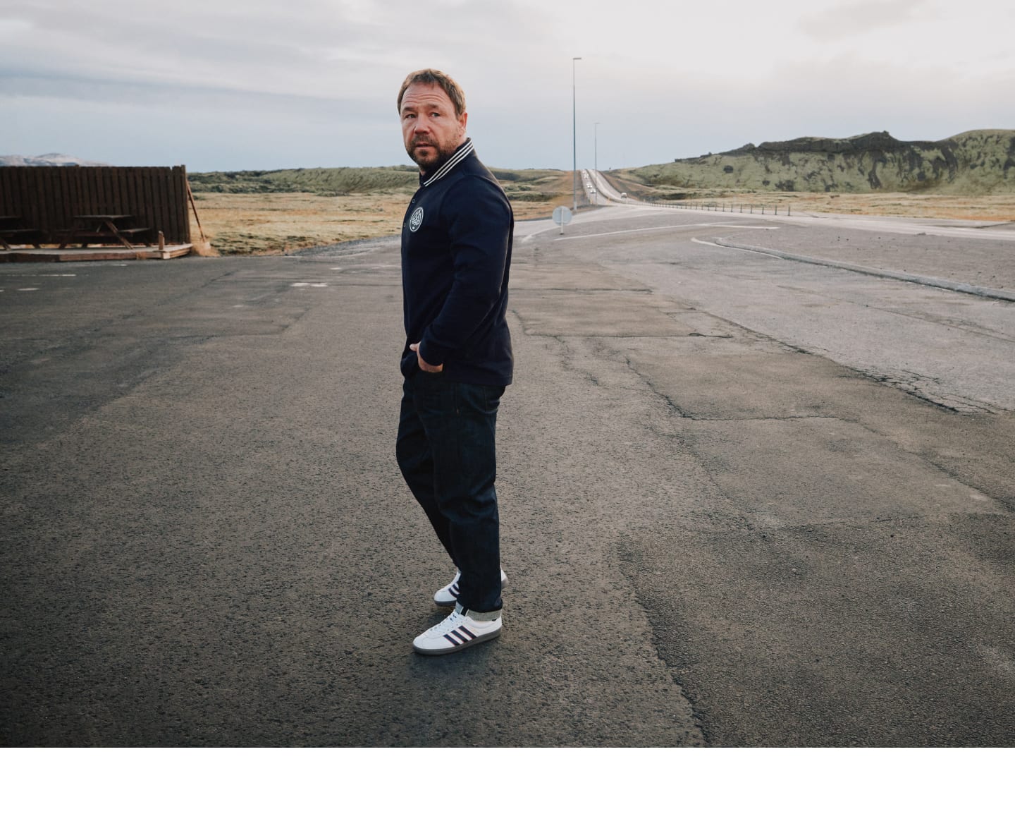 Stephen Graham walks on an asphalt road surrounded by empty fields, wearing a black jacket from the 2023 adidas Spezial collection.
