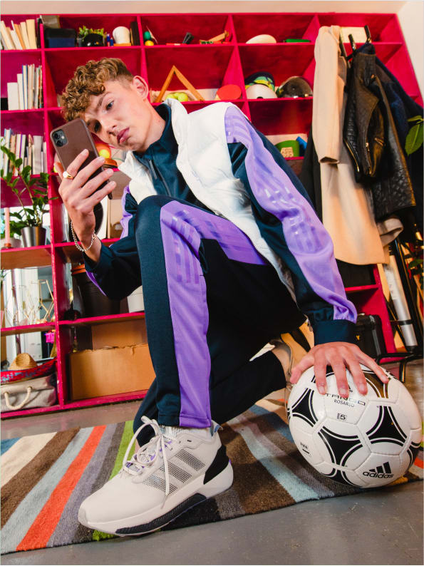 Young man wearing adidas tracksuit poses in squat for cell phone selfie with soccer ball.