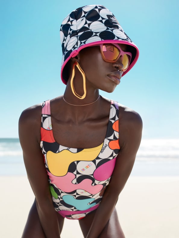 A female model poses on a beach, styled in bucket hat and swimsuit from the adidas x Rich Mnisi Pride Collection.