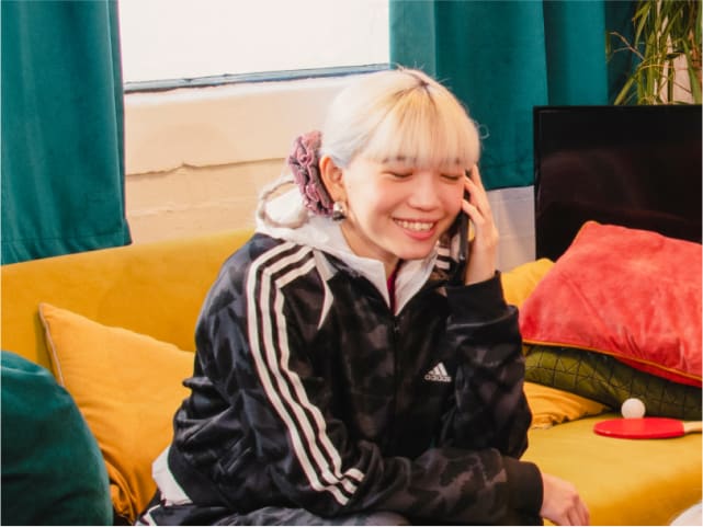 Smiling girl wearing adidas tracksuit and hoodie sits on couch with phone in hand.
