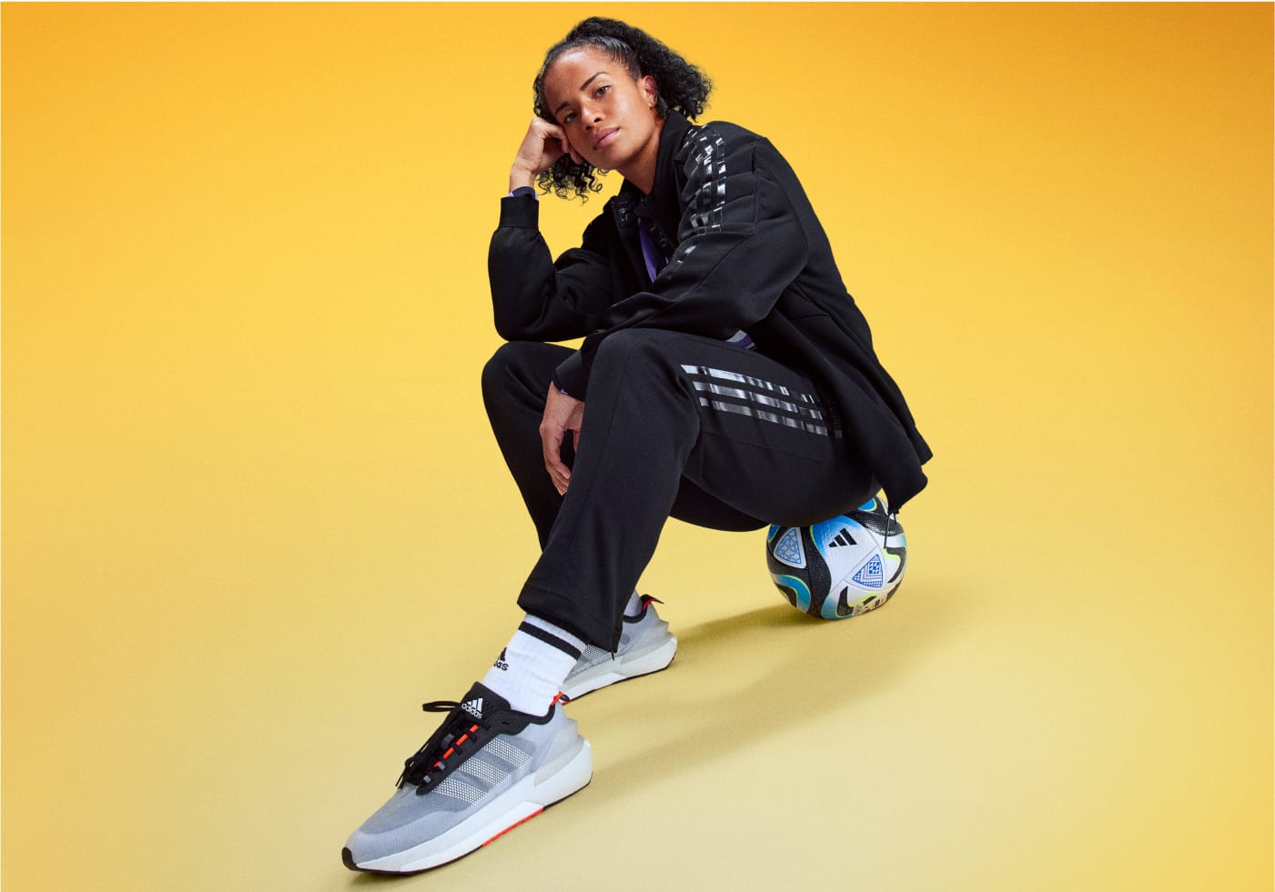 Soccer player Mary Fowler sits posed on a soccer ball, wearing a black adidas tracksuit and sneakers.