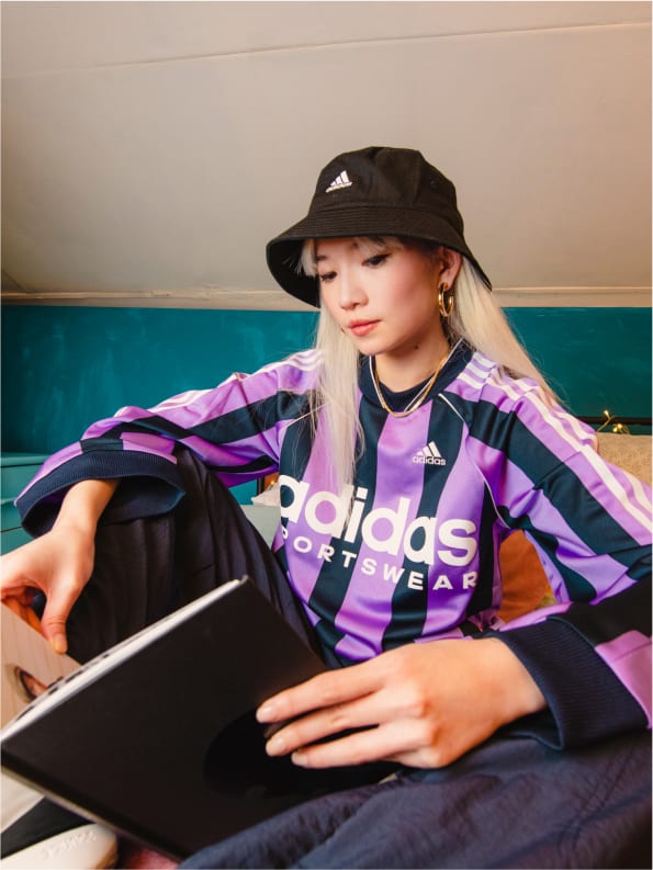 Girl wearing adidas jersey and bucket hat seated with book open on couch.