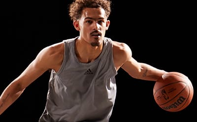 A waist-up view of professional basketball player Trae Young dribbling with his left hand in front of a black background