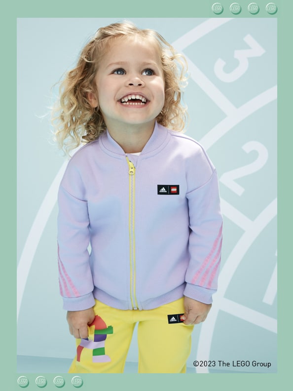 An infant girl standing in front of a printed athletics track, smiling playfully, wearing adidas LEGO® onesie.