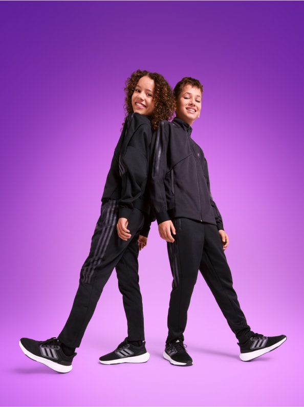 Two children, one male one female, stand smiling back to back in black adidas tracksuits and sneakers.