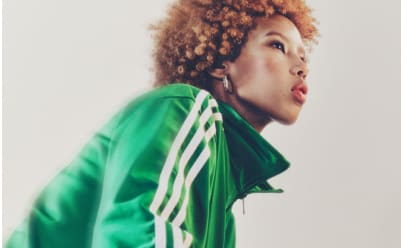 A portrait of a woman with red hair wearing a green adicolor tracksuit and golden earrings and standing in front of a white backdrop.