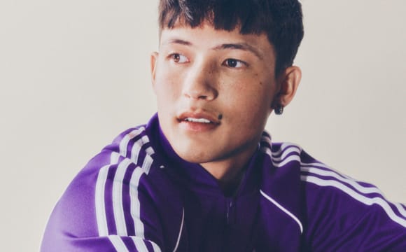 A portrait of a man with black hair wearing a purple adicolor tracksuit in front of a white backdrop.