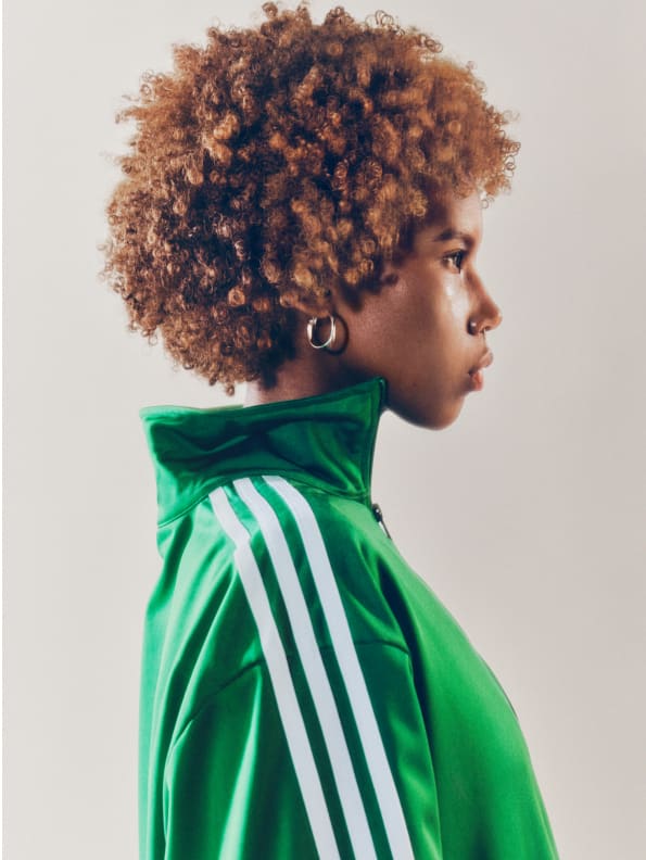 A portrait of a woman with red hair wearing a green adicolor tracksuit and golden earrings and standing in front of a white backdrop.