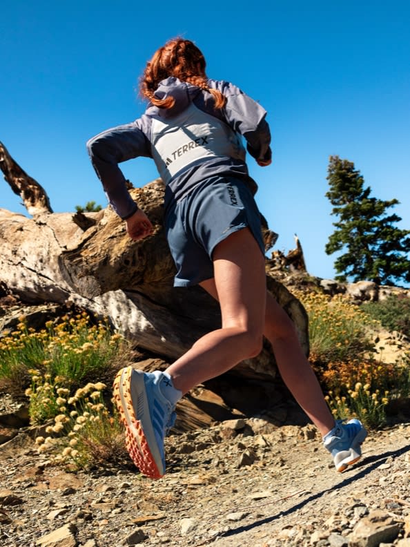 Female running in the mountain view from the side wearing Wearing the TERREX SOULSTRIDE FLOW trail running shoes, the Windweave jacket and TERREX apparel.