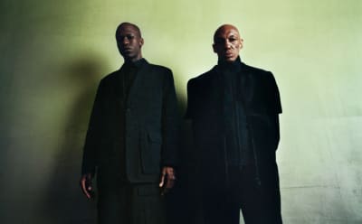 Two men in black Y-3 apparel stand in front of a green backdrop and stare blankly into the camera.