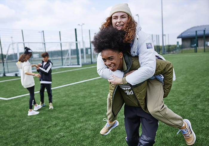 Man gives woman a piggyback ride on a sports field with two children playing in the background.