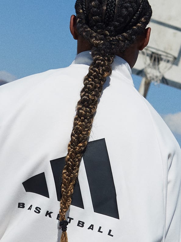 A rear view of a person with long braided hair dribbling a ball and wearing a white sweatshirt with an adidas Basketball logo