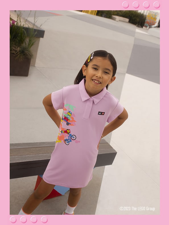 A little girl standing in front of a bench, smiling confidently wearing adidas LEGO® dress and footwear