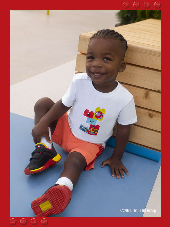 An infant boy sitting by a bench, smiling confidently wearing adidas LEGO® t-shirt, shorts and footwear.