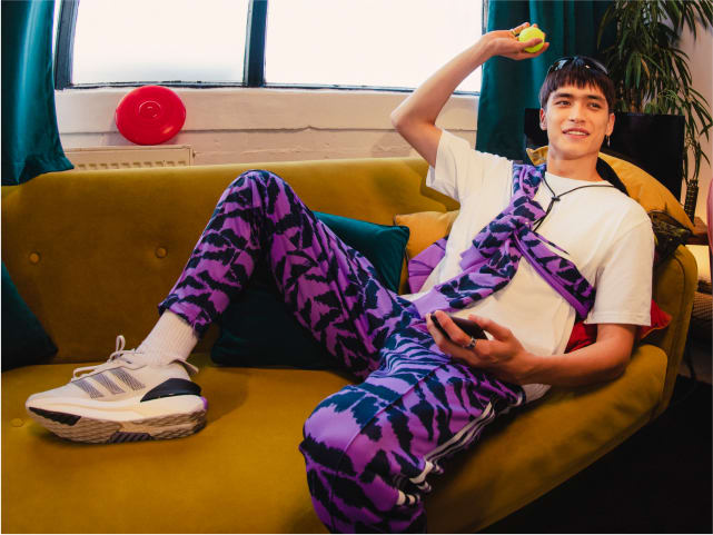 Boy dressed in adidas tracksuit and sneakers lounges casually on couch with tennis ball and phone in either hand.