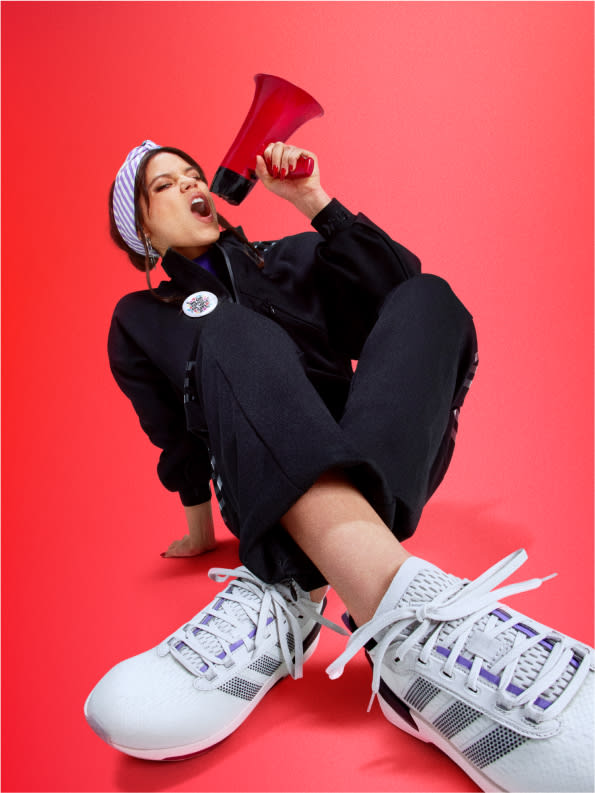 Actress Jenna Ortega sits on the ground yelling into a megaphone enthusiastically, wearing adidas tracksuit and sneakers.