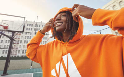 WNBA player Erica Wheeler smiles as she lifts her orange adidas hoody away from her face. She's standing on a basketball court, with city buildings behind her.
