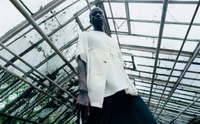 A man stands in a greenhouse, wearing a white Y-3 shirt and black pants while staring down at the camera.