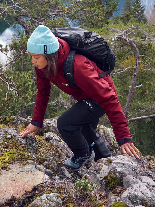 Women wears a red Terrex Xperior gore-tex paclite rain jacket with a blue hat and a black backpack whilst climbing over some rocks against a backdrop of trees.