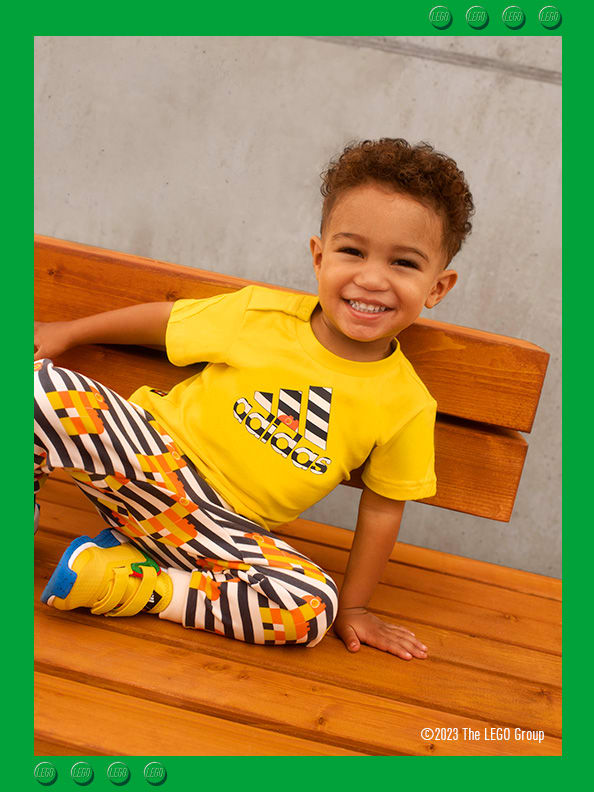 A little boy sitting on a brick floor, smiling confidently wearing adidas LEGO® t-shirt, trousers and footwear.