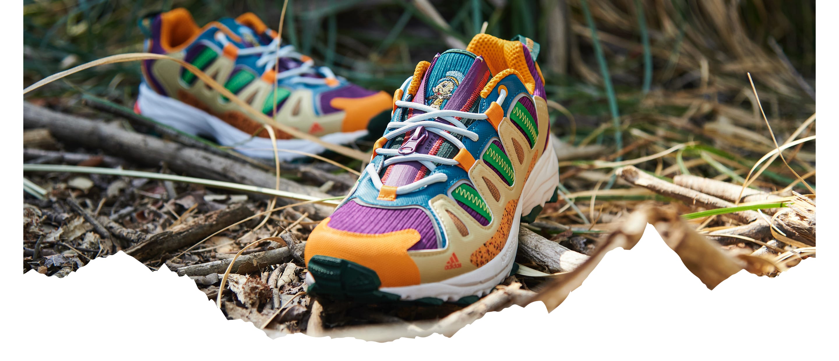 A pair of colourful Superturf Adventure sneakers on the ground, covered with sticks, branches and grass.