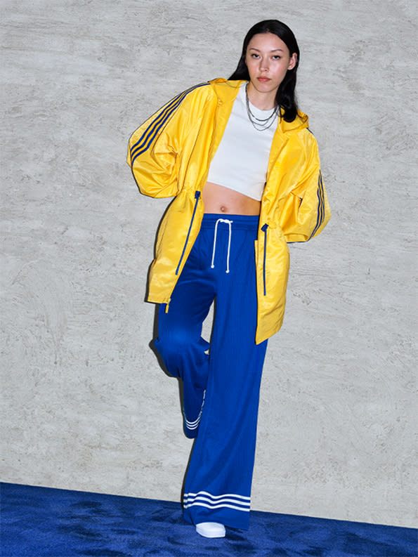 A model wearing blue trackpants and a yellow jacket from the latest adicolor collection with Gazelle shoes, standing on blue carpet against a grey concrete wall