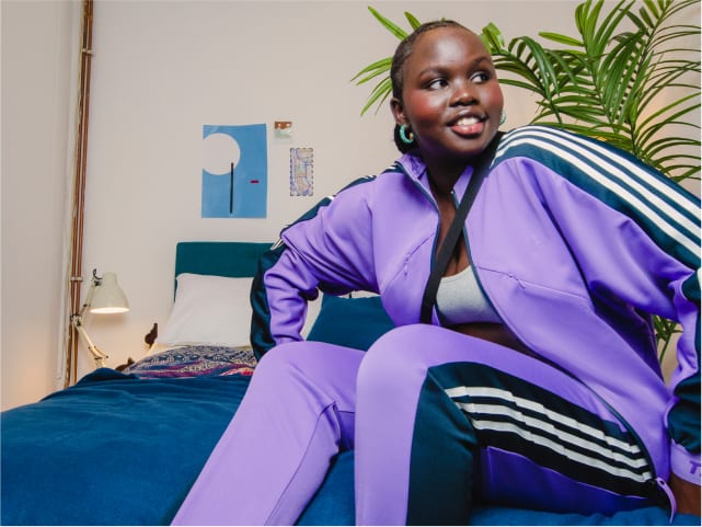 Girl dressed in purple adidas tracksuit sits smiling on bed.