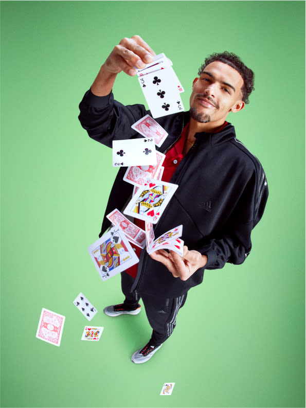 Basketball player Trae Young, dressed in black adidas tracksuit and sneakers, shuffles a deck of cards.