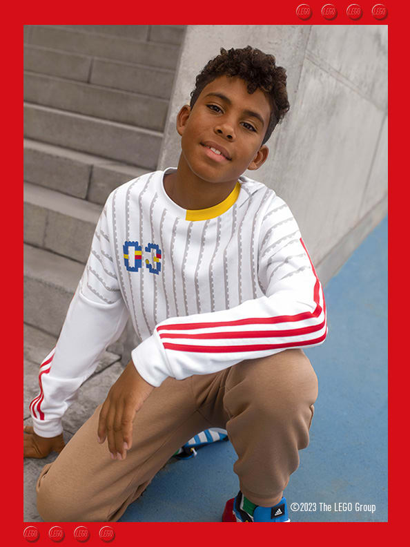 A young boy crouching next to concrete steps, smiling confidently wearing adidas LEGO® sweatshirt, trousers and footwear.