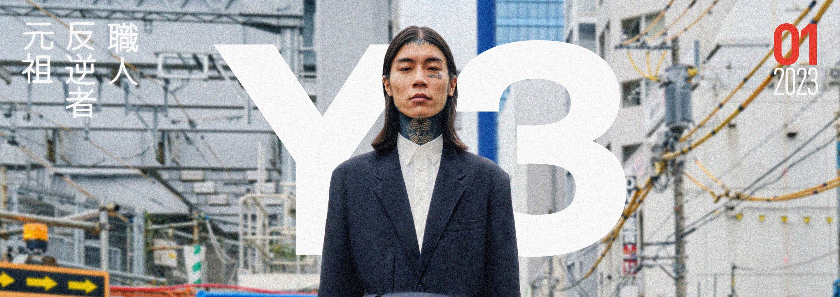A man with long black hair and neck tattoos stares into the camera, standing on a busy street while wearing a black suit and a white shirt.