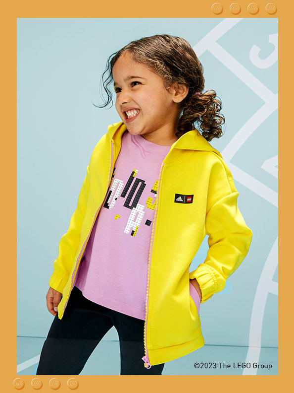 A little girl standing in front of a printed athletics track, smiling playfully with one hand in her pocket, wearing adidas LEGO® jacket, tee and trousers.