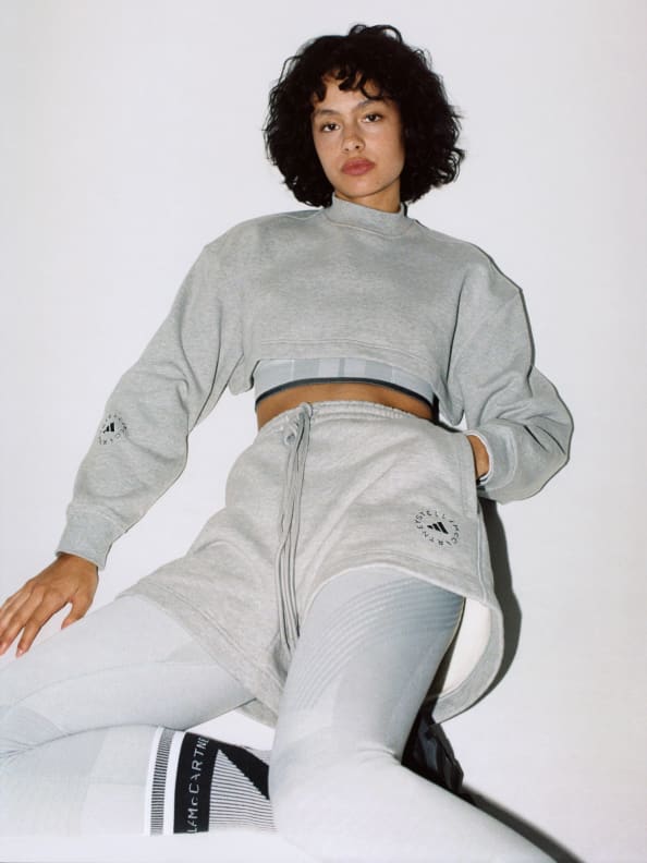 A model sitting on the floor. She wears a layered look of the cropped sweatshirt with cropped T-shirt underneath on top. On bottom she wears the grey tight underneath the Terry shorts.