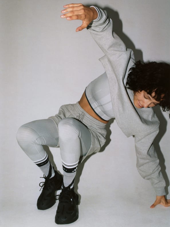 A model poses dynamically on the floor. She wears a layered look of the cropped sweatshirt with cropped T-shirt underneath on top. On bottom she wears the grey tight underneath the Terry shorts.