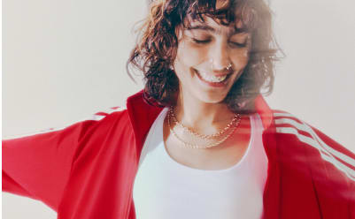 A portrait of a woman with brown hair wearing a red adicolor tracksuit in front of a white backdrop.