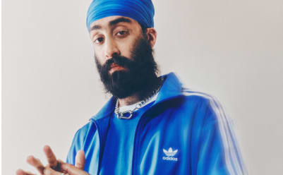 A portrait of a man in a blue turban and a blue adicolor tracksuit standing in front of a white backdrop.