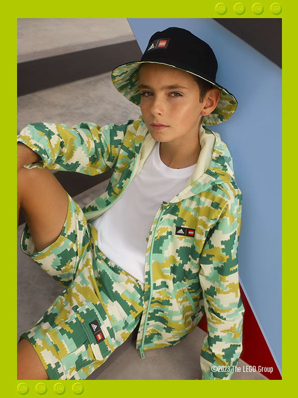 A young boy sitting confidently on concrete steps, wearing adidas LEGO® bucket hat, hoodie, t-shirt and shorts.