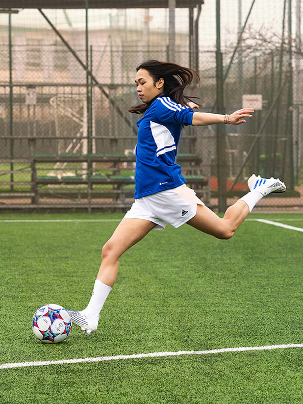 Image of female player, blue and white kit, kicking the ball on the pitch.