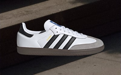 A pair of white and black Adidas sneakers rests on a dark surface, with a bright light casting a shadow across the image.